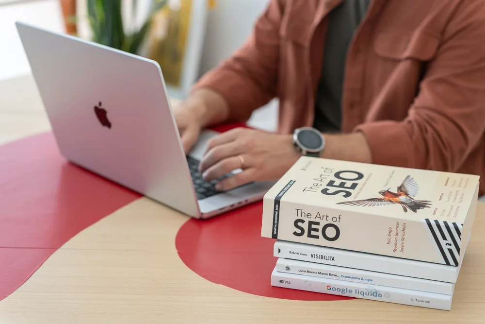 Dettaglio delle mani sul portatile accanto alla pila di manuali di SEO